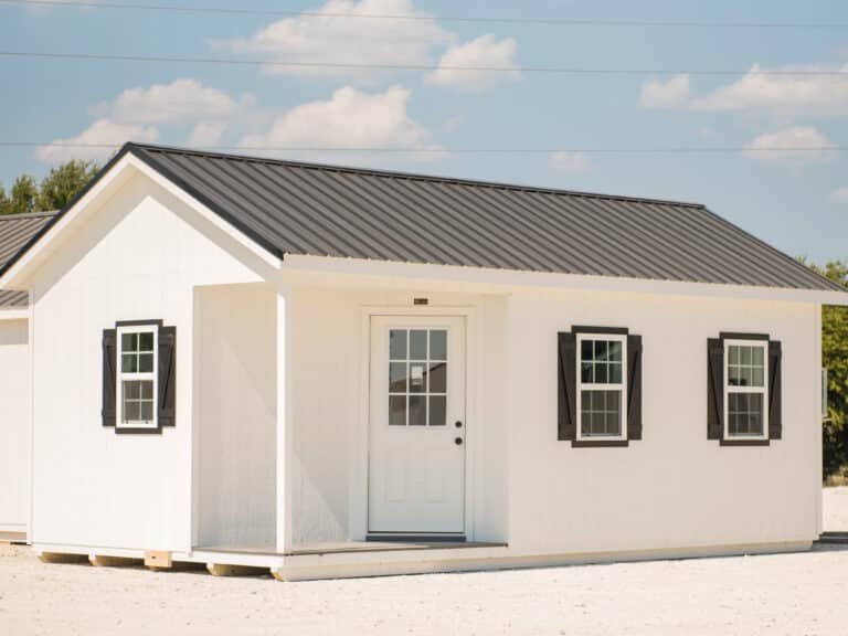 White Outpost cabin with black roof and trim, displayed on a gravel lot under a bright Texas sky.