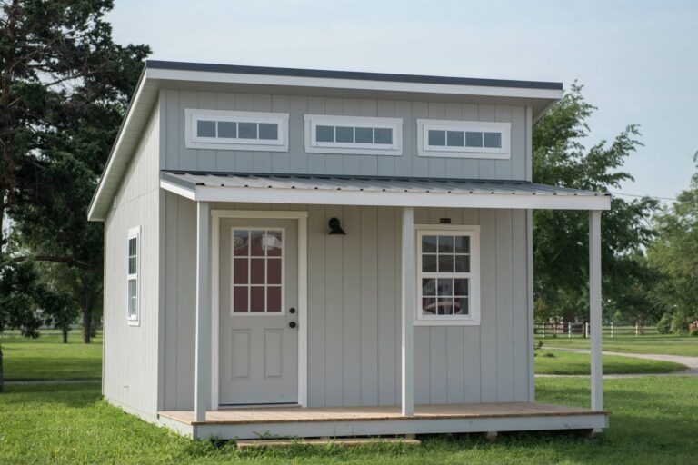 Light gray modern portable cabin with front porch and clerestory windows, placed on a grassy lot.