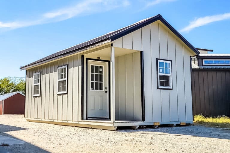 Beige portable Outpost cabin with black trim, white door, and windows, displayed on a gravel lot under a bright blue Texas sky.