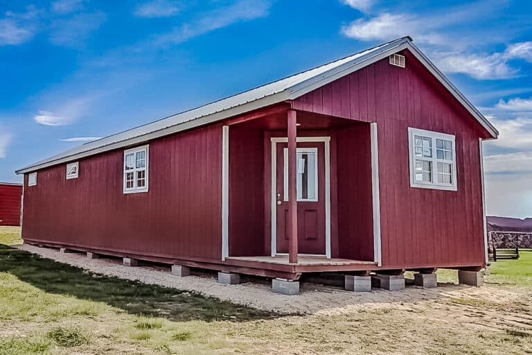 Red Outpost cabin with white trim and a small covered porch, resting on concrete blocks in a grassy yard beneath a clear blue sky.